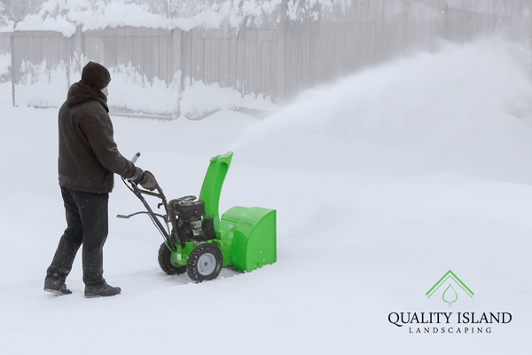A man covered in snow gear pushing a green snow plow with snow blowing towards the fence and the matching quality island green logo